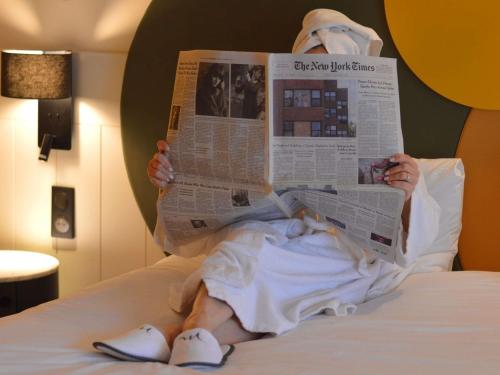 a person laying on a bed reading a newspaper at Mercure Auxerre Autoroute du Soleil in Appoigny