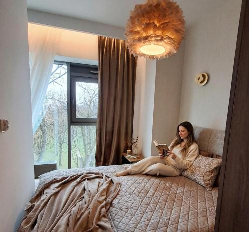 a woman sitting on a bed reading a book at Park View Olszynki Park in Rzeszów
