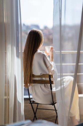 a woman sitting in a chair holding a cup of coffee looking out a window at Guest House Velik in Sandanski