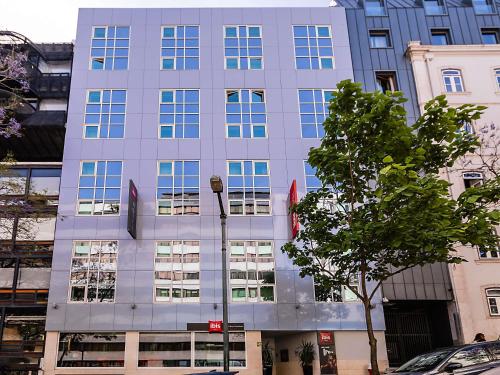 a tall gray building with a tree in front of it at Hotel ibis Lisboa Liberdade in Lisbon