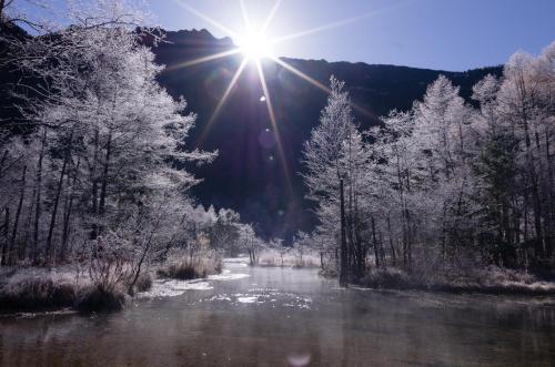 a river with snow covered trees and the sun in the sky at Kamikochi Lemeiesta Hotel in Matsumoto