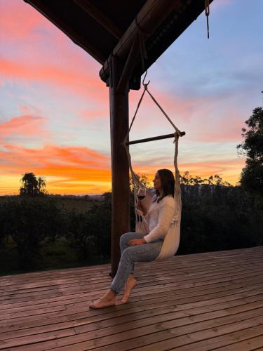 a woman sitting on a porch swing at sunset at Chalé romântico com jacuzzi e vista do pôr-do-sol in Harmonia