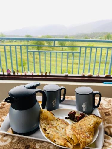 a plate of food on a table with two mugs and food at Aahista Stays- Mountain view homestay in Bīr