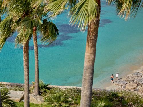two people walking on a beach with palm trees at Mykonos Blu, a Resort to Live in Psarou