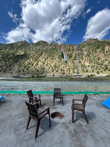 three chairs and an umbrella on a beach with a river at Kishan Ganga Camping Site in Wāmpor
