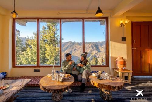 a man and woman sitting in a room with large windows at Zostel Homes Gadagushaini, Jibhi in Jibhi