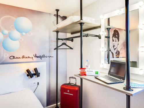 a bedroom with a desk with a laptop and a red suitcase at Ibis Styles Paris Saint Denis La Plaine in Saint-Denis