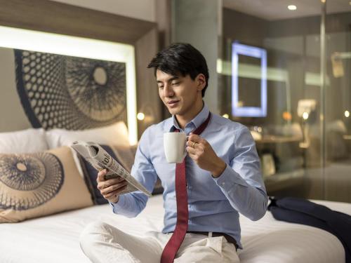 a man sitting on a bed holding a coffee cup and a newspaper at Novotel Bangkok Bangna in Bangna