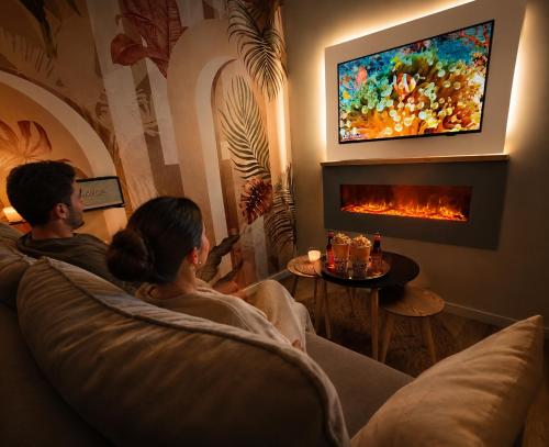 a couple sitting on a couch in a living room with a fireplace at MRODBNB - Appart Neuf - Aperçu Mer - 25m Plage & Resto - Ciné - cheminée - Popcorn in Cancale