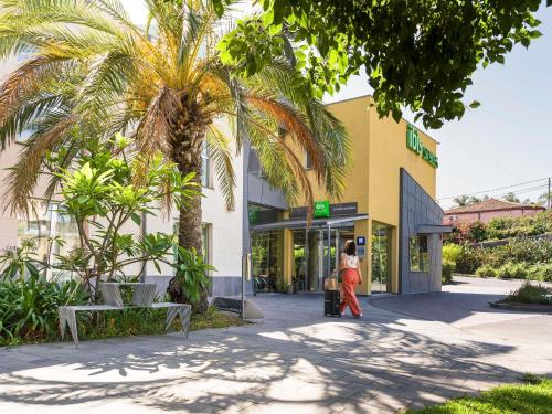 Eine Frau geht vor einem Gebäude mit einer Palme entlang. in der Unterkunft ibis Styles Catania Acireale in Acireale