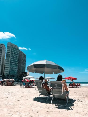 two people sitting in chairs under an umbrella on the beach at Grand Hotel Guarujá - A sua Melhor Experiência Beira Mar na Praia! in Guarujá