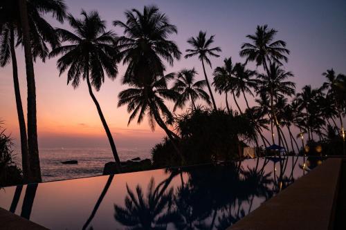 a pool with palm trees and the ocean at sunset at Kumu Beach By Teardrop Hotels in Balapitiya