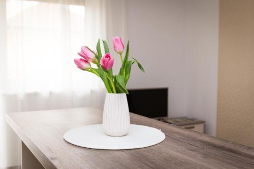 a white vase with pink flowers in it on a table at Aqualiget-Apartmanház E-Car plug-in point Szeged in Szeged