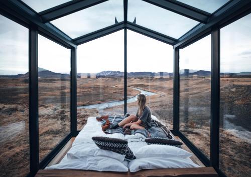 a woman sitting on a bed in a room with large windows at Panorama Glass Lodge SOUTH in Hella