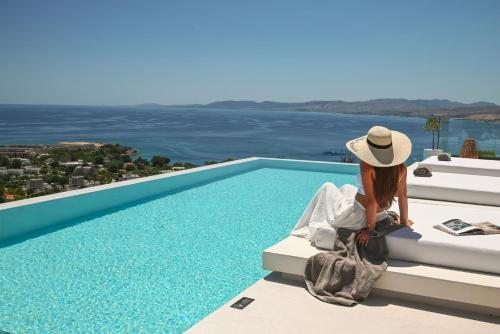 a woman sitting on a ledge next to a swimming pool at Aethra Villa - Infinity Pool & Panoramic Sea View in Pefki