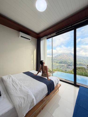 Eine Frau sitzt auf einem Bett in einem Zimmer mit einem großen Fenster in der Unterkunft Pinggan Infinity - Private Pool Villa in Kintamani in Gretek