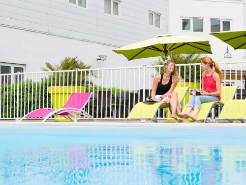 two women sitting under umbrellas next to a swimming pool at Novotel Paris Orly Rungis Aéroport in Rungis