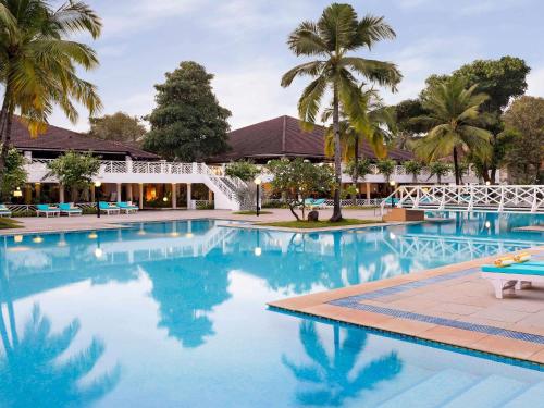 a large swimming pool with palm trees and a white bridge at Novotel Goa Dona Sylvia Resort in Cavelossim