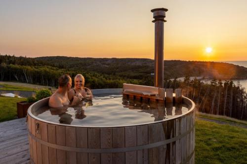 two people sitting in a hot tub with the sunset at Birch - Luxury Oceanview Cottage with Hot Tub & Private Beach Access near Cabot Trail in Chimney Corner