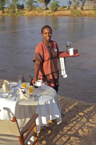 a man standing next to a table in front of a river at Ashnil Samburu Camp in Archers Post