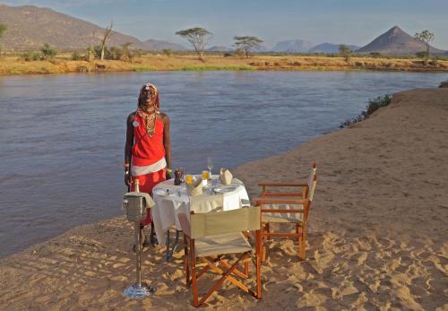 a woman standing on the shore of a river with a table at Ashnil Samburu Camp in Archers Post