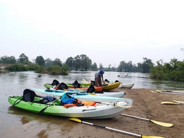 Several colorful kayaks lined up on a sandy riverbank with a person preparing one kayak under overcast sky