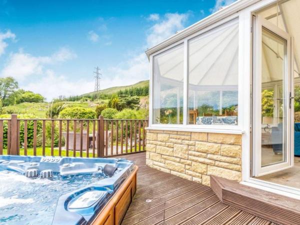 Outdoor hot tub on wooden deck next to glass conservatory with green hills and blue sky in background