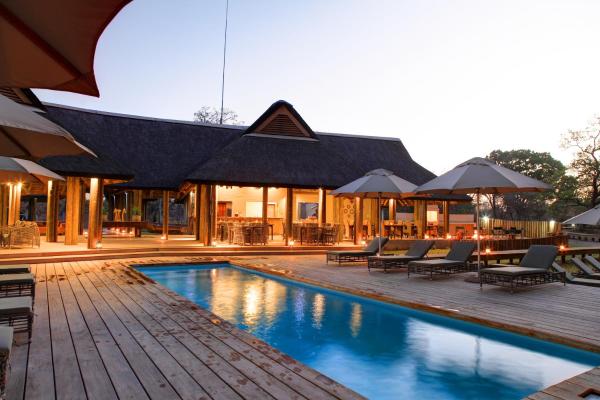 Outdoor pool with lounge chairs and umbrellas at dusk beside a thatched-roof building with warm lights