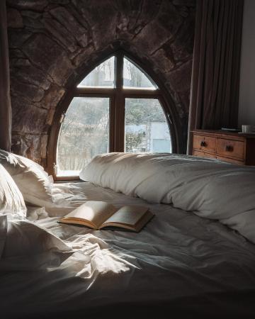 bed with white linens and open book near arched stone window with wooden frame