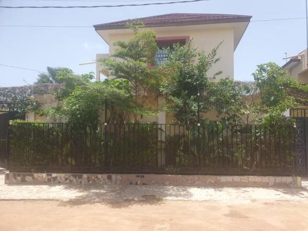 two-story building behind black metal fence with dense green trees and plants in front under clear sky