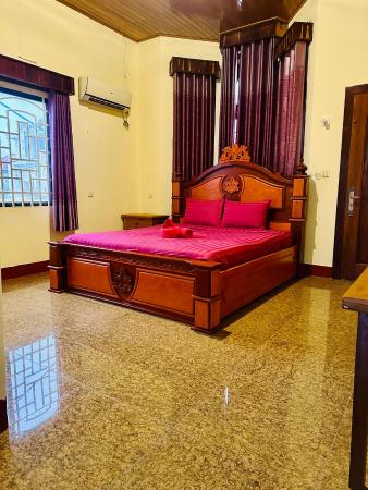 Room with wooden bed frame, red bedding, air conditioner, window with bars and curtains, and tiled floor