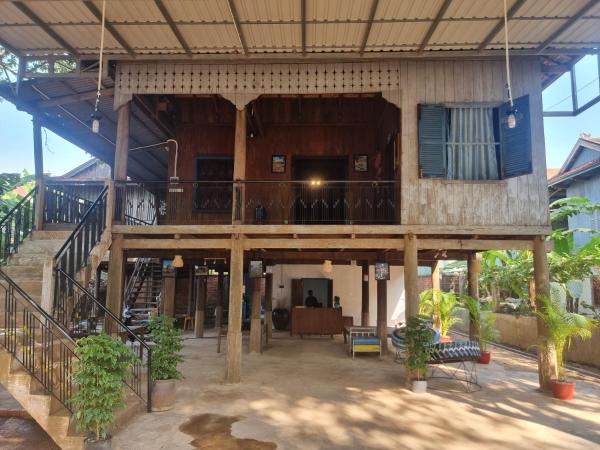 Traditional wooden stilt house with open ground floor, stairs, potted plants, and seating area under natural daylight