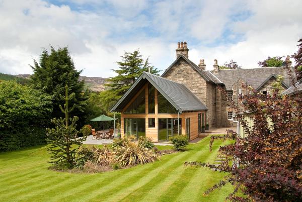 Stone house with modern wooden extension surrounded by green lawn, trees, and outdoor seating under umbrella