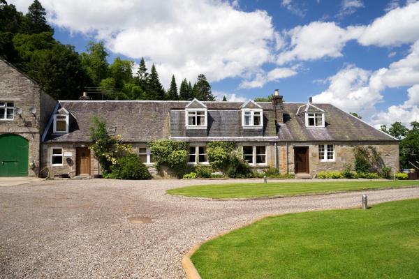 Long stone cottage with dormer windows, green lawn, gravel driveway, and trees under a blue sky with clouds